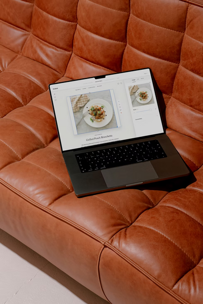 Laptop on a brown leather sofa displaying a website with food images.
